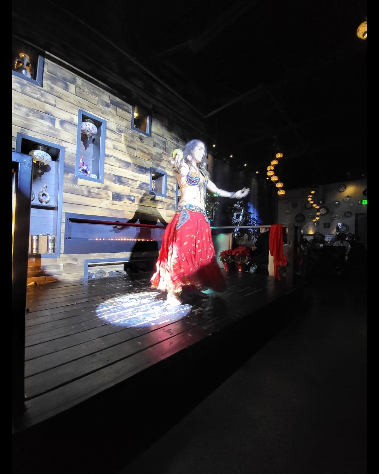 A woman is performing traditional dance on a stage in a dimly lit restaurant or bar with wooden decor and hanging lanterns in the background.