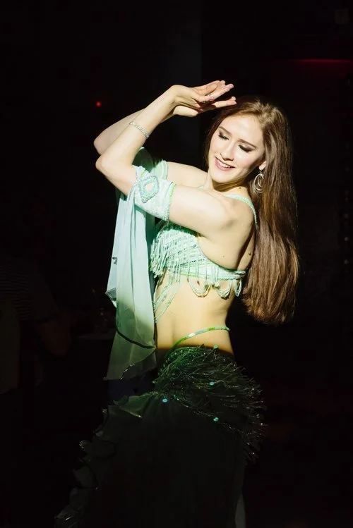 Young woman dancing in a dark setting, wearing a green crop top and black skirt with feathers, smiling with eyes closed.