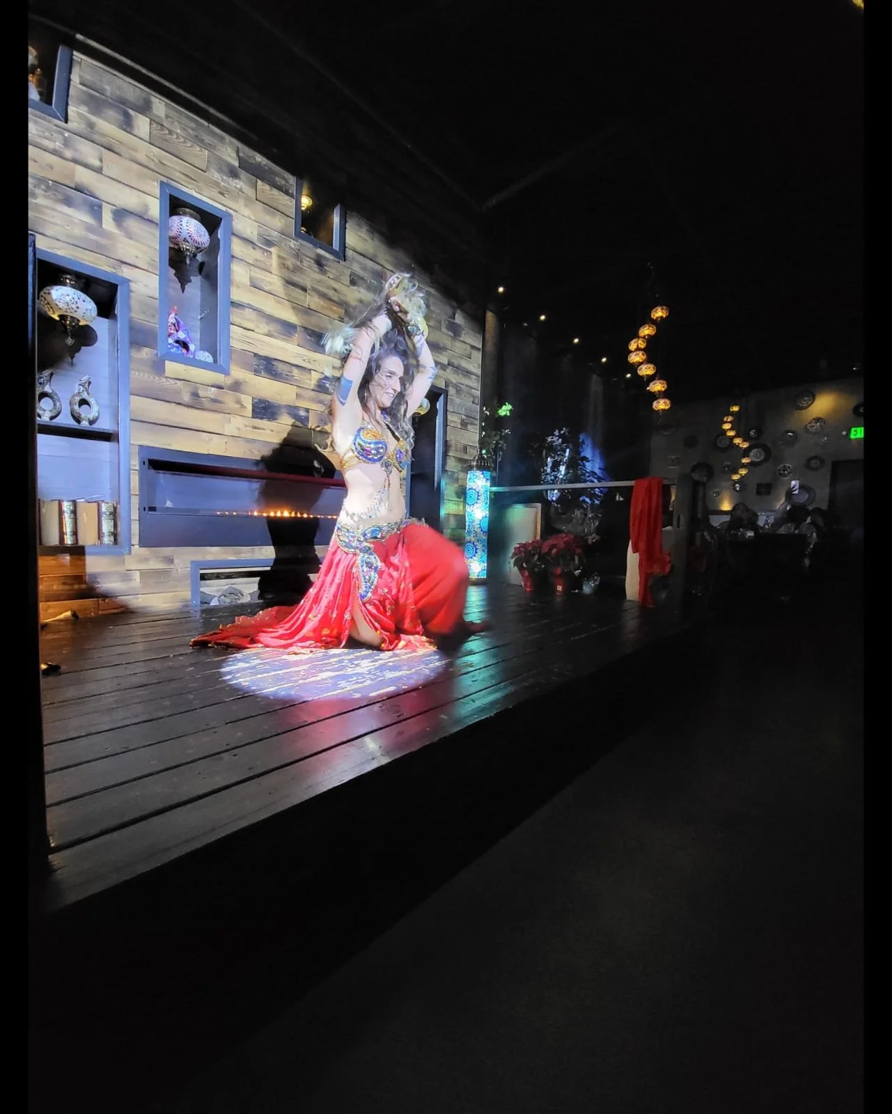A woman performing a belly dance on a stage with wooden floor and decorative background, wearing a red skirt and decorated top, holding a veil or scarf above her head, with stage lighting and hanging decorations in the background.