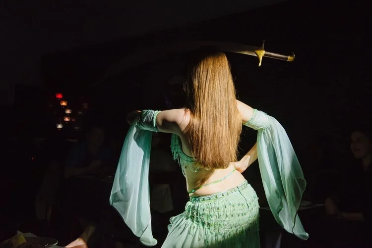 A woman with long red hair dressed in a belly dance costume holding a sword on her shoulder, performing in a dimly lit setting.
