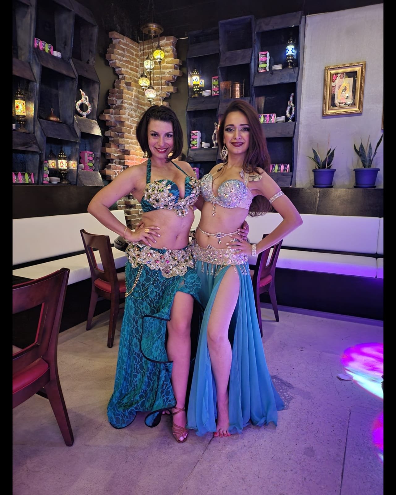 Two women in colorful, embellished costumes posing inside a restaurant or lounge with dark wooden shelves, brick accent wall, and potted plants.
