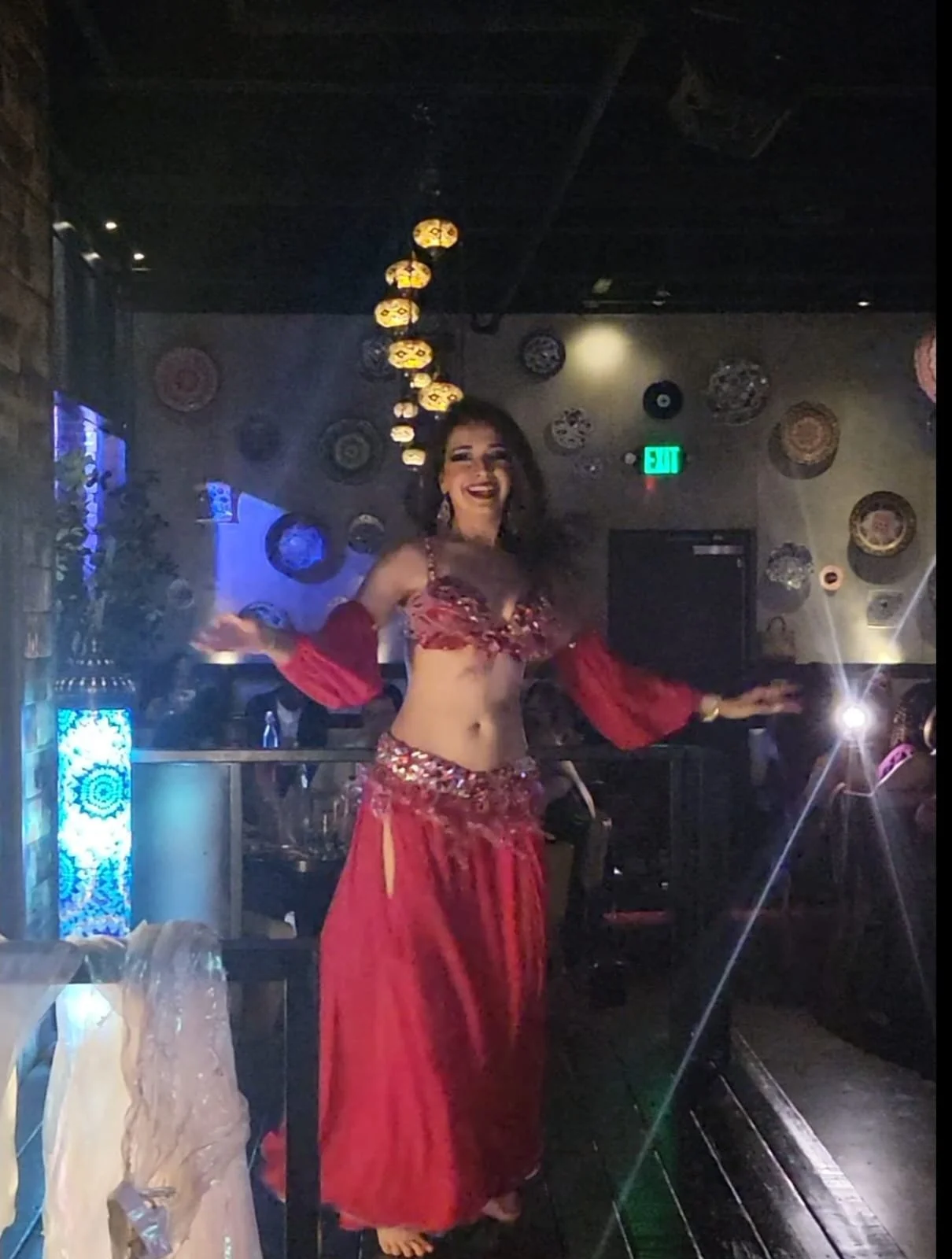 A woman dancing in a red belly dancing costume with a cropped top and long skirt inside a dimly lit venue decorated with colorful circular wall art and hanging lanterns, smiling happily.