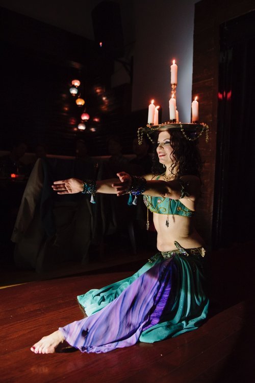 A woman in a colorful belly dance costume performs with a candle crown on her head during a dark, festive event.