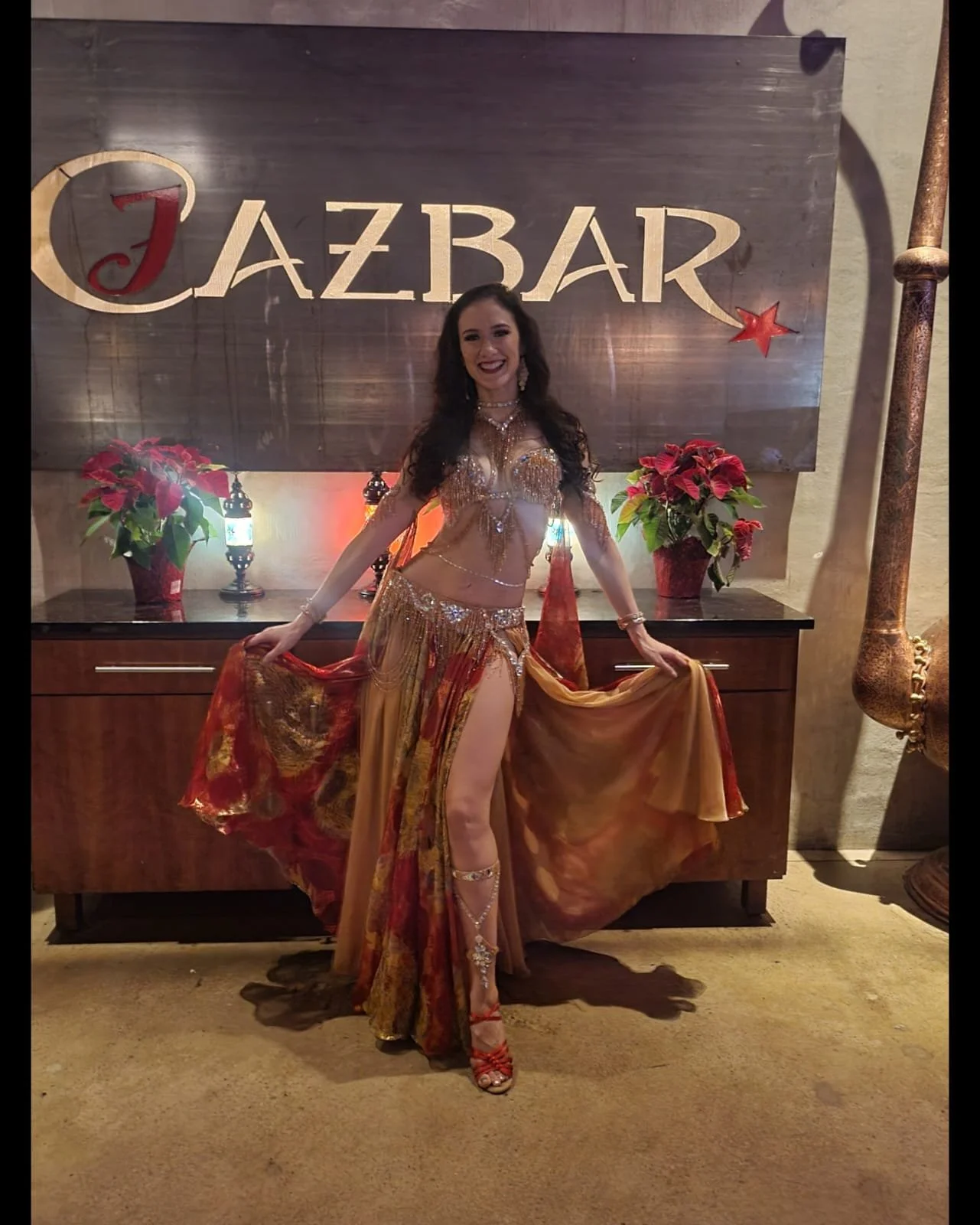 A woman in a gold and red belly dancing costume standing in front of a sign that says "Jazzbar." She is smiling and posing with her arms out, holding the flowing sides of her costume. There are red and green poinsettia plants on a dark wooden table b