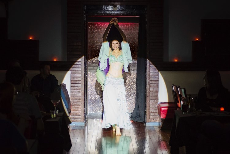 A woman in belly dance costume standing in doorway on a stage or runway, with people seated at tables watching in a dimly lit room.