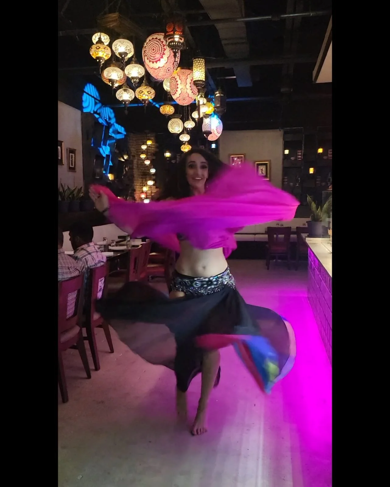 A woman dancing in a dimly lit restaurant or bar with colorful hanging lanterns. She is spinning with a pink fabric in her hands, wearing a black skirt with a colorful train and a glittery top. She has long curly hair and a big smile.