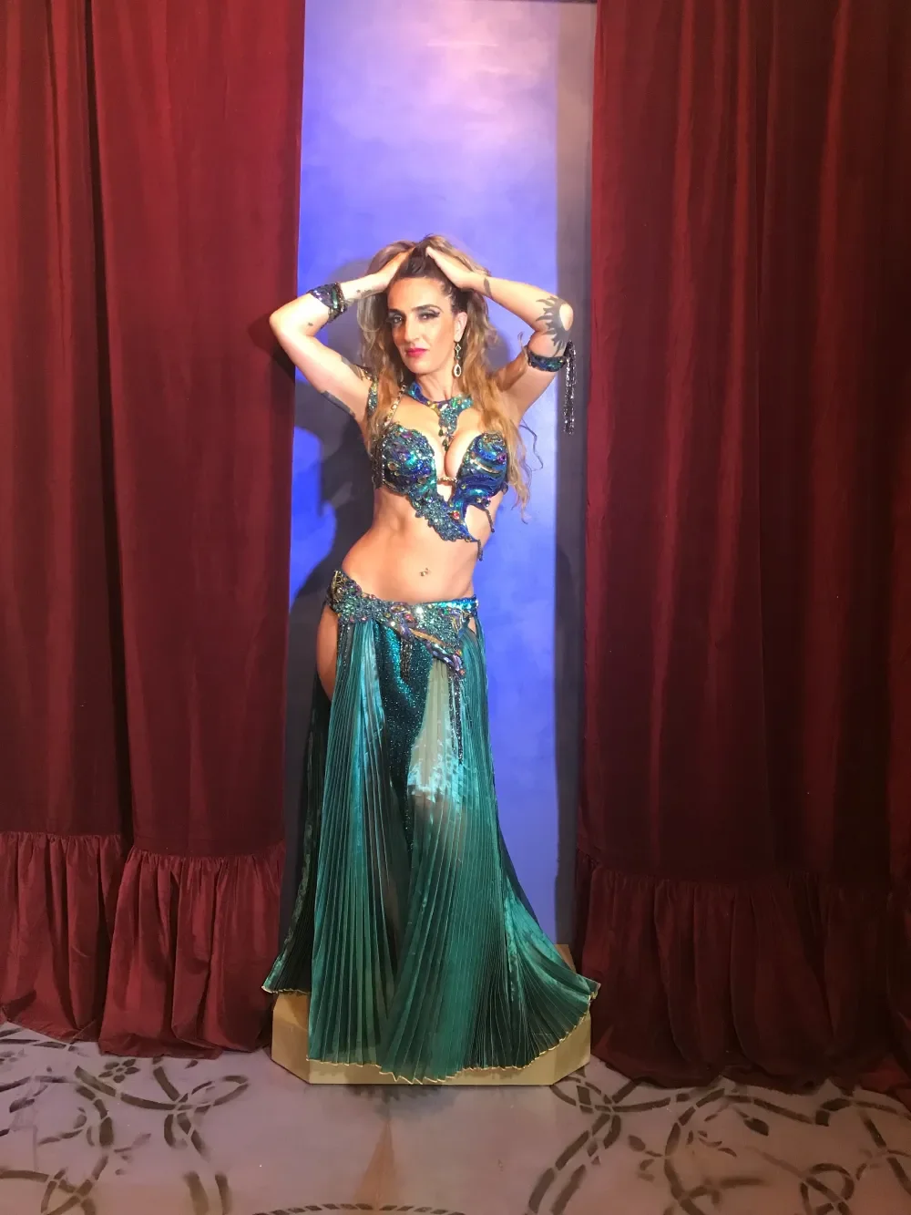 Woman dressed in a turquoise and blue belly dance costume standing between red velvet curtains.
