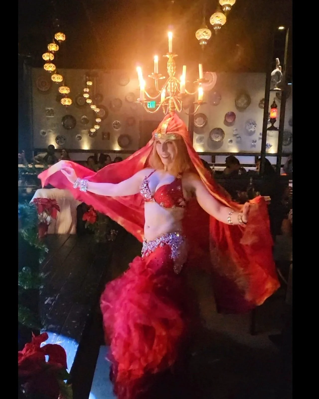 A performer dressed in a red and gold belly dance costume with a red cape, performing indoors under a lit chandelier with decorative wall plates in the background.