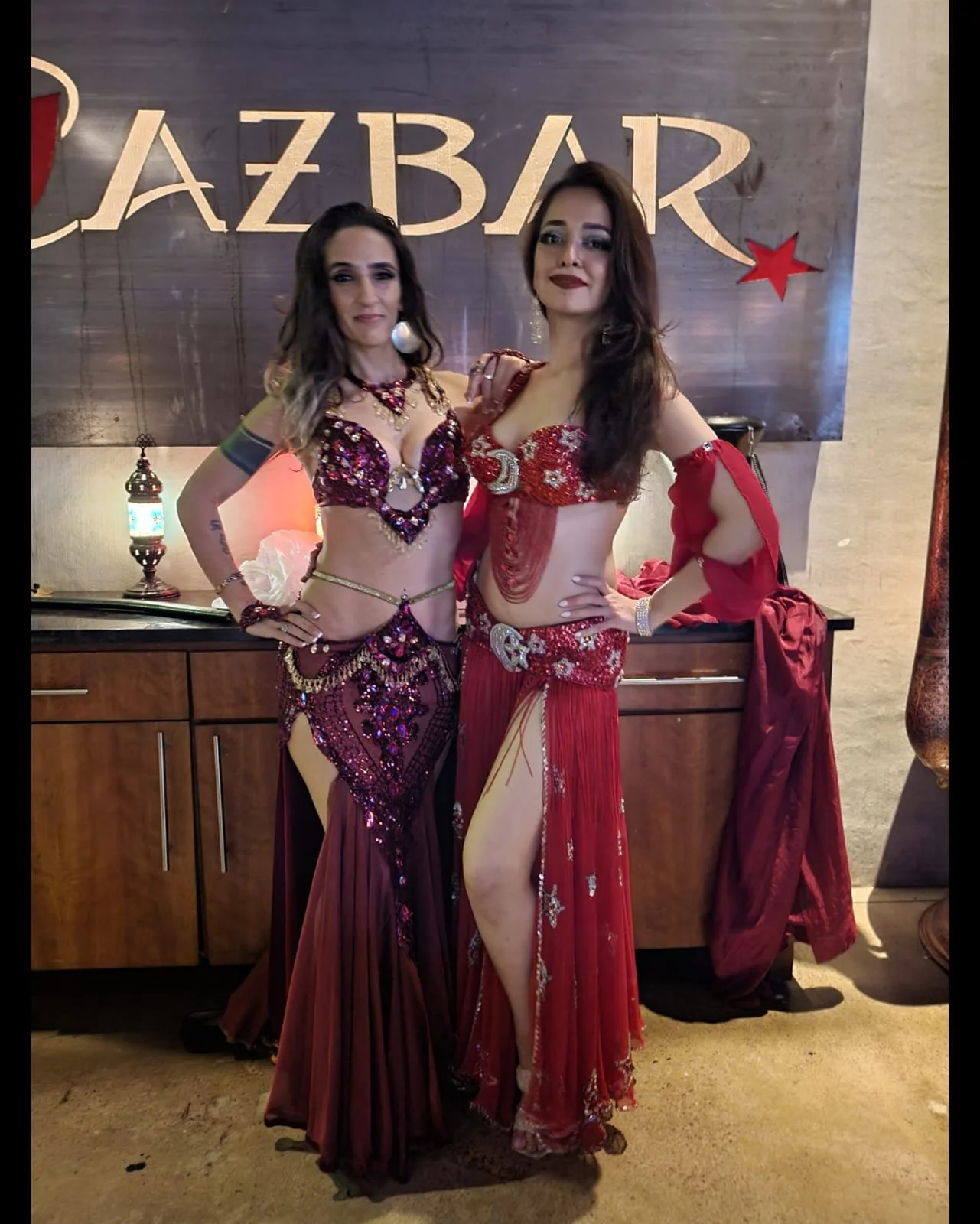 Two women in elaborate belly dance costumes standing in front of a wooden backdrop with the word 'JAZZBAR' and star decorations, posing for a photo.