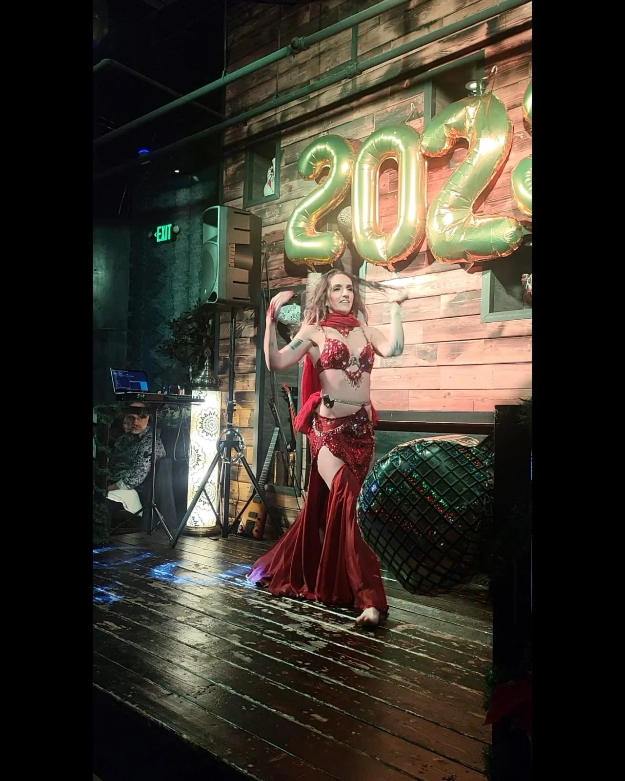 A woman in a red, glittery belly dance costume performing on stage with large gold balloons shaped as the numbers 2022 in the background.
