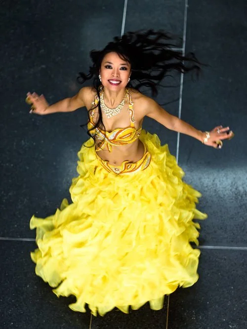 A woman in a yellow flamenco-style dress, smiling and spinning in mid-air, with dark hair flowing, on a black tiled floor.