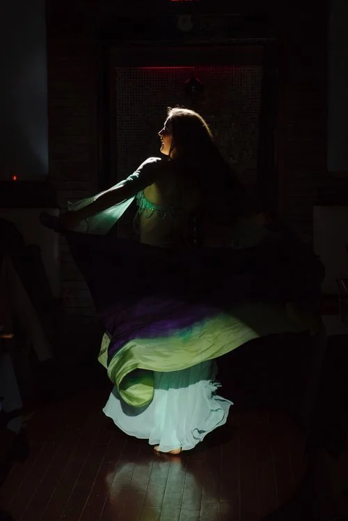 A woman with long hair wearing a flowing dress is spinning in a dimly lit room, with light illuminating her silhouette from the side.