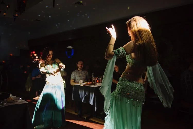 Two women in belly dance costumes performing for a small audience in a dimly lit room.