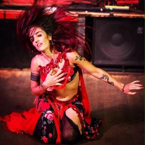 A woman dressed in a red and black costume, dancing on stage with her hair flying and a smile on her face.