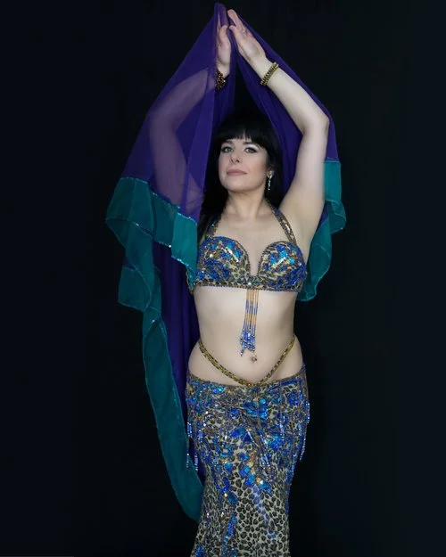 Woman in a colorful belly dance costume with a purple and turquoise veil, posing with arms above her head against a black background.
