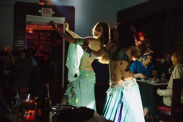 Two women in belly dance costumes perform in a dimly lit restaurant with people seated at tables in the background.