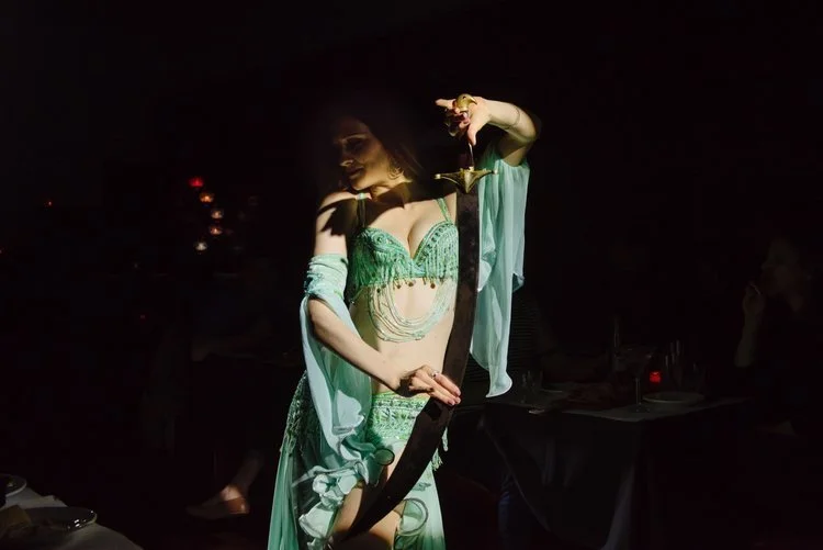 A woman in a green and gold belly dance costume holding a curved sword in a dimly lit room.