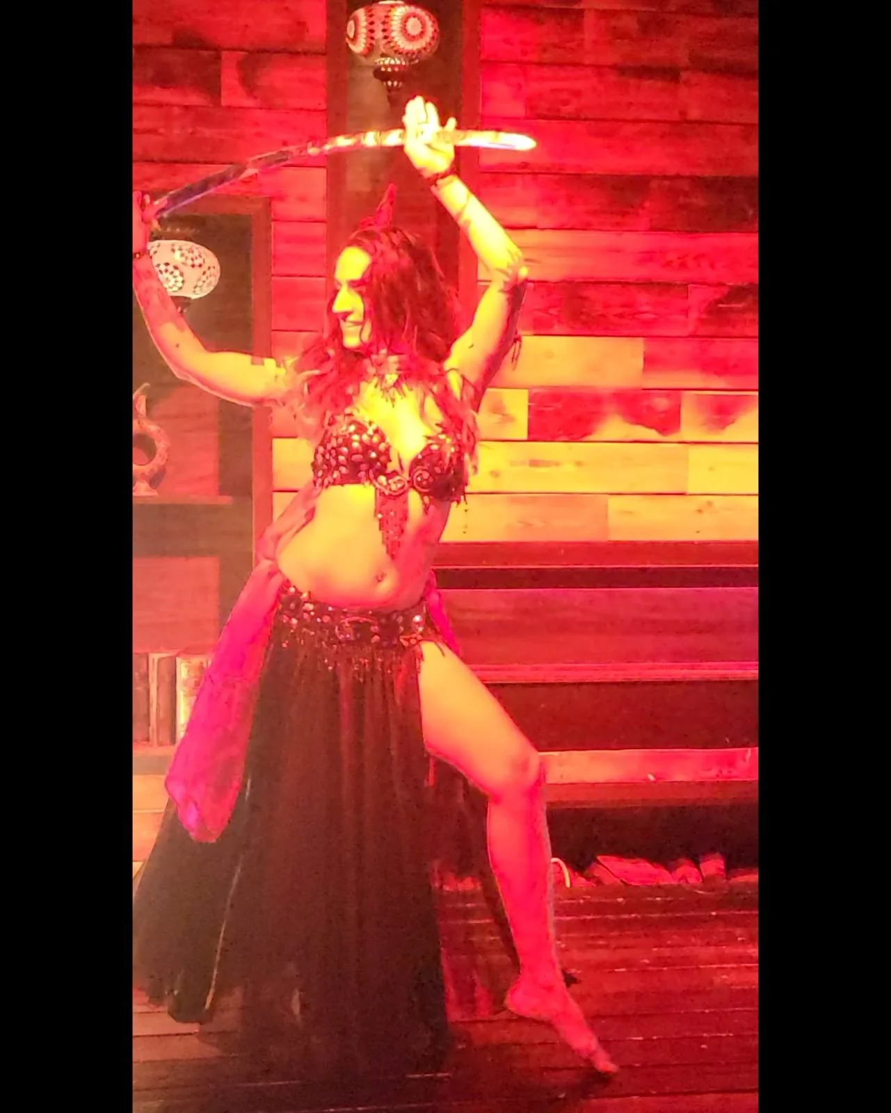 A woman dressed in a belly dance costume with a black skirt and red top, posing with a sword and holding a lit ring above her head, in front of a wooden wall with hanging lamps.