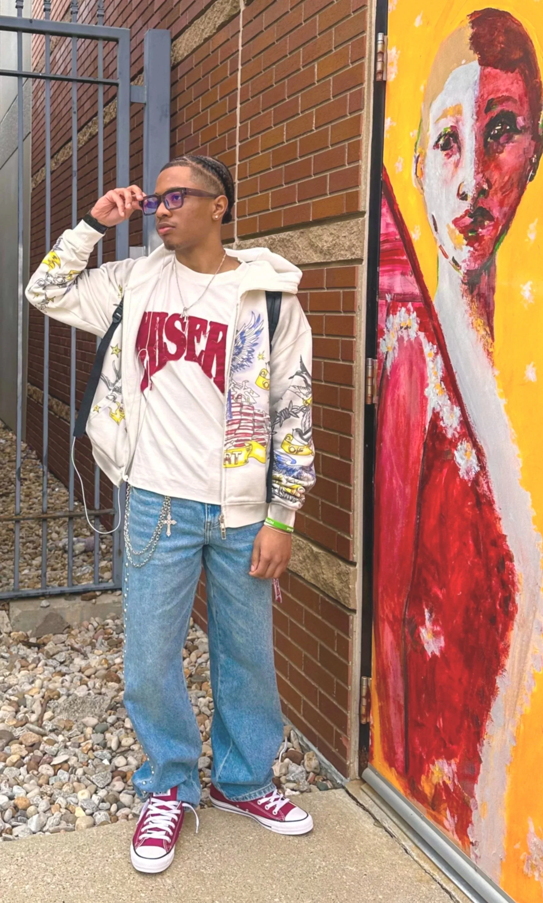 Young person wearing glasses, white hoodie with colorful designs, jeans with chain, red sneakers, standing outdoors on gravel next to an abstract portrait painting of a woman with yellow background.