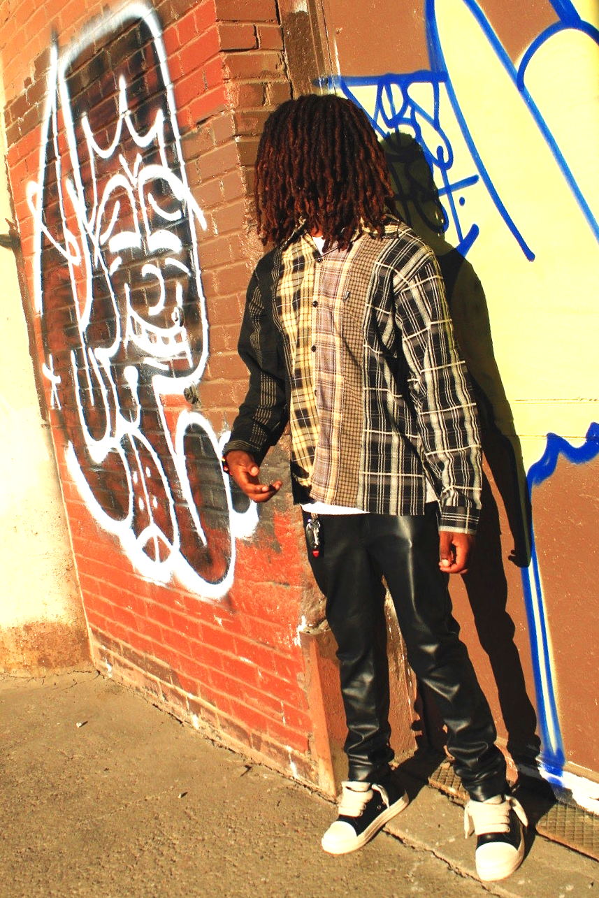 A person with dreadlocks stands in front of a graffiti-covered brick wall, wearing a mixed plaid shirt, black leather pants, and sneakers, casting a shadow.