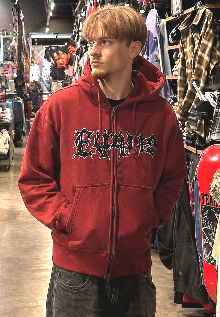 A young man with light brown hair and facial hair wearing a red hoodie with black and white text, standing in a store aisle filled with clothing and accessories.