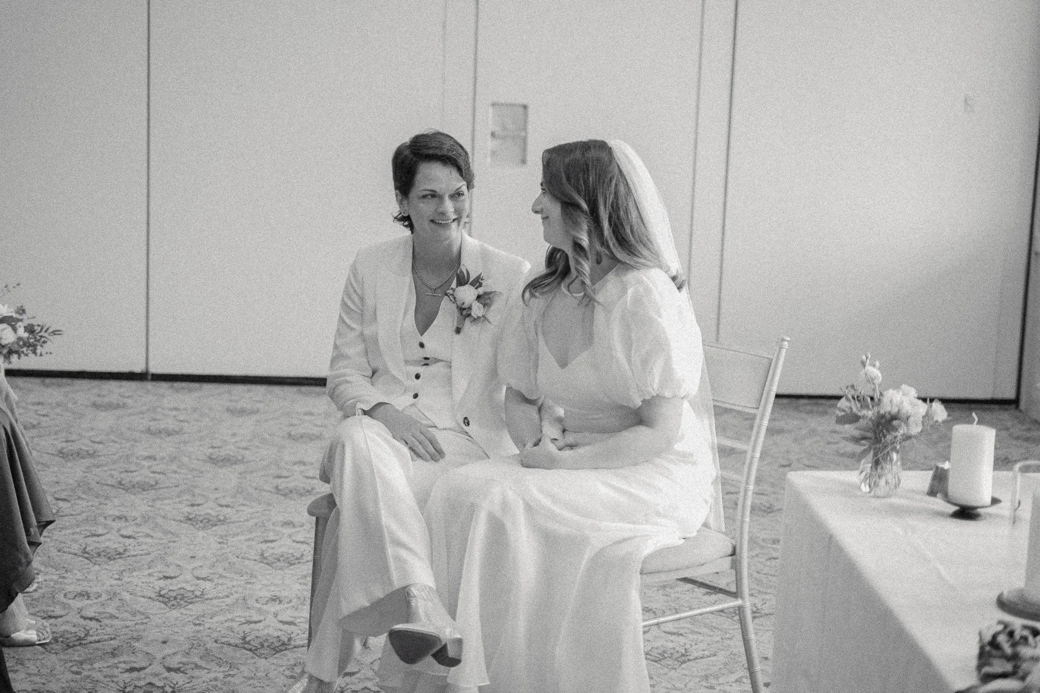 Two women, possibly a bride and her officiant or friend, engaged in conversation at a wedding ceremony in a simple indoor setting. Both women are seated on chairs, with the woman on the right wearing a wedding dress and veil, and the woman on the lef