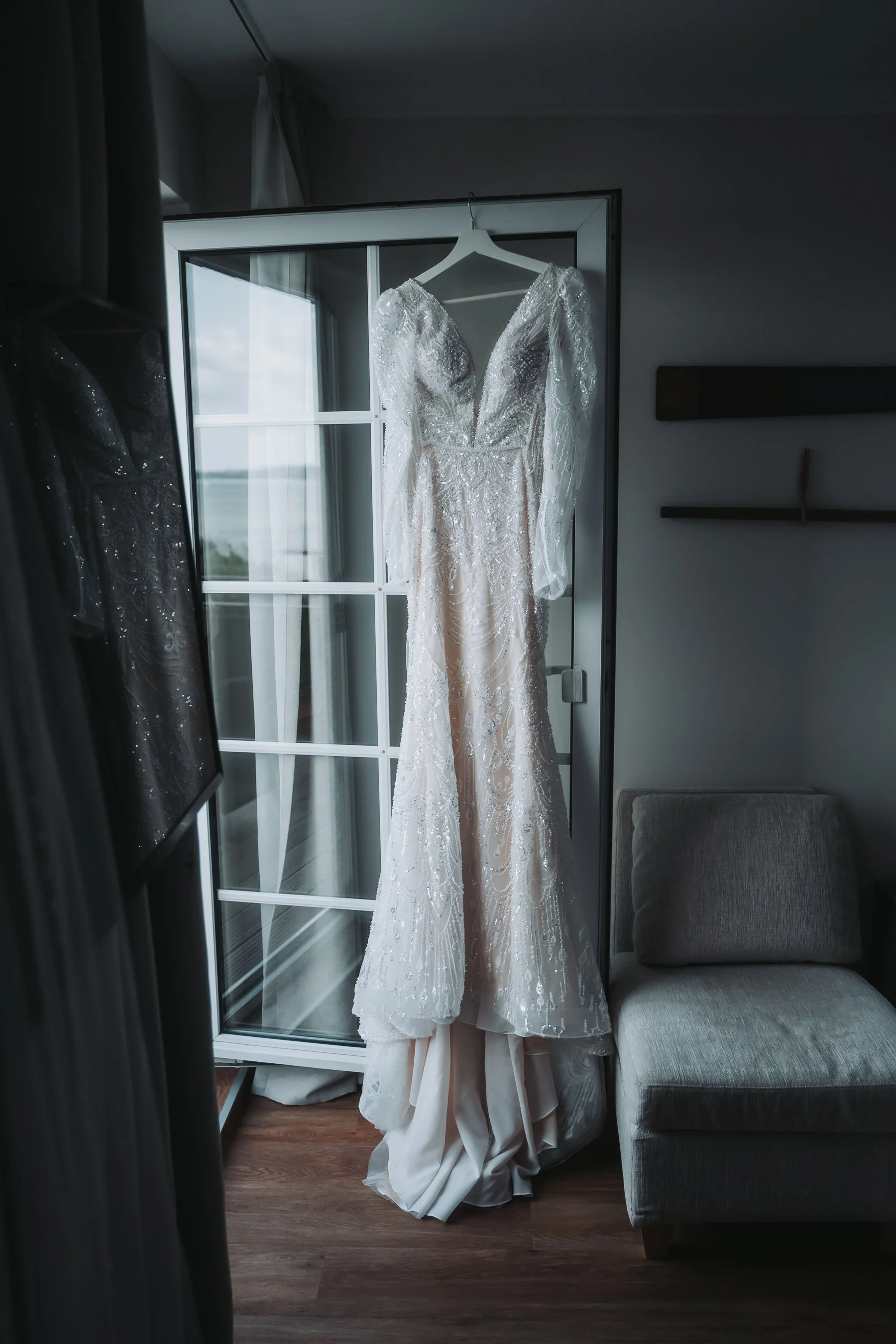 A wedding dress with intricate beading and lace details hanging on a hanger in front of a glass door, with a mirror and curtains reflecting part of the room.