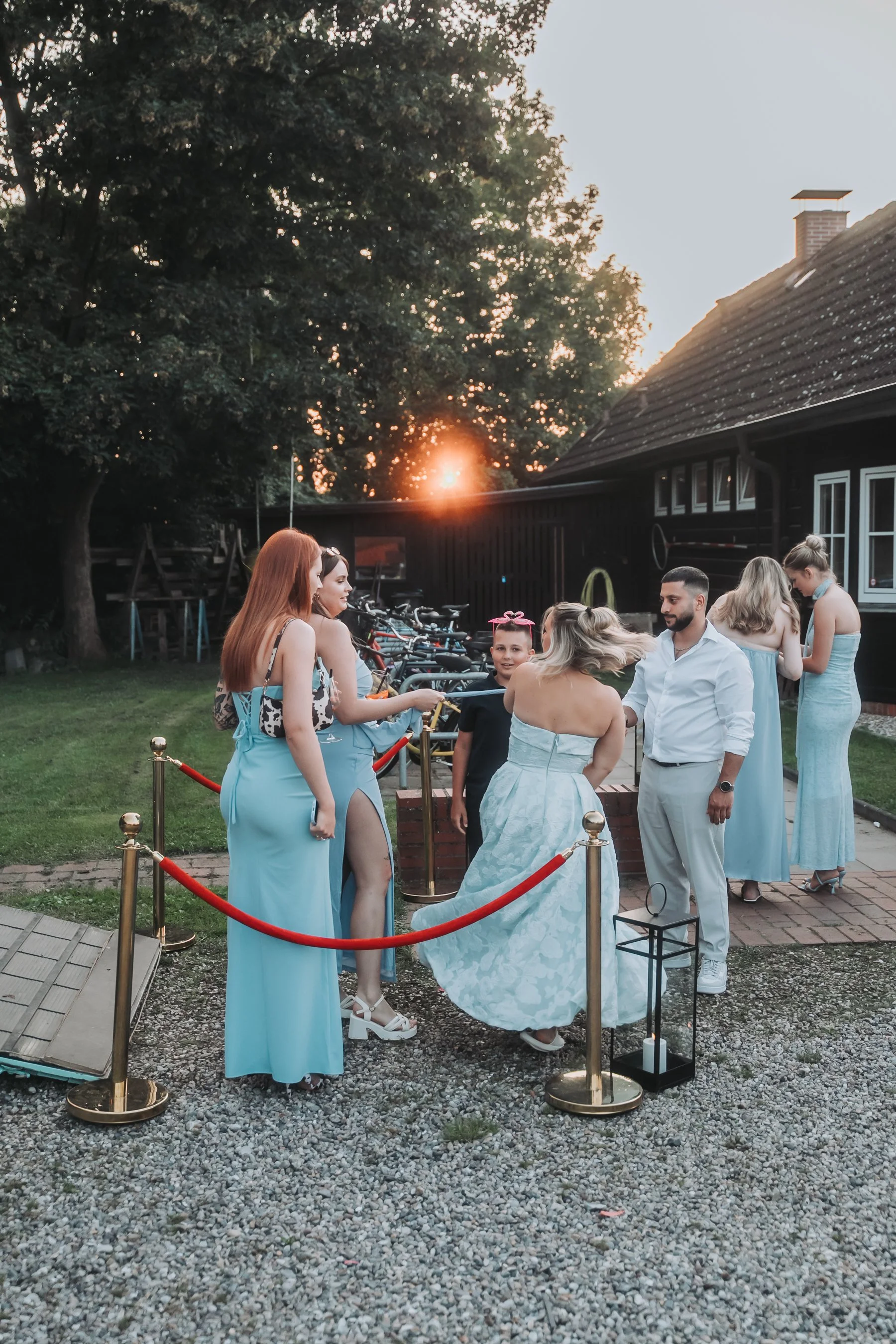People dressed in formal attire, including long dresses and suits, are gathered outdoors at sunset. They are standing behind a red rope barrier, with bicycles lined up in the background nearby. A lantern and a lantern-like decoration are on the groun