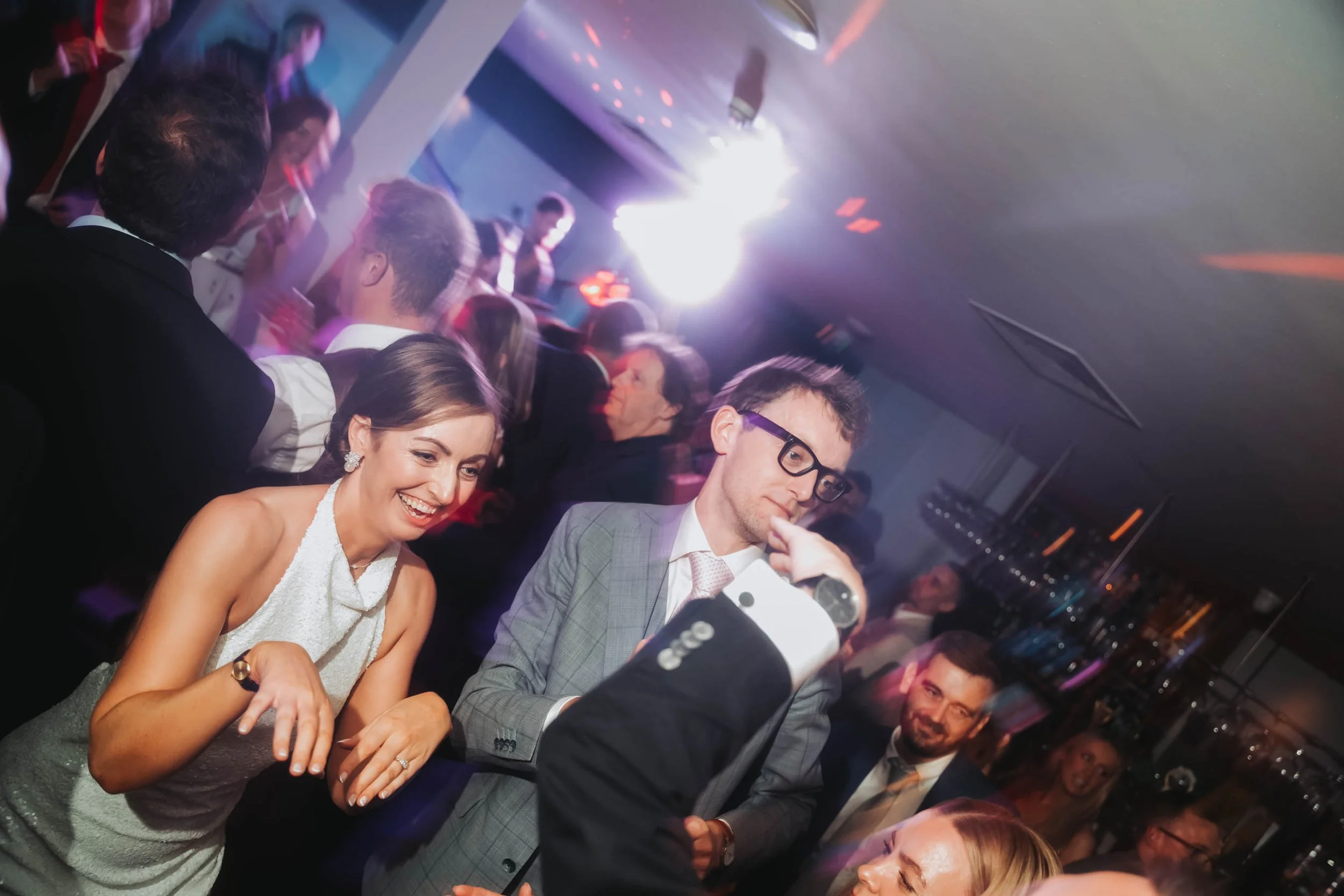 People dressed in formal attire dancing and socializing at a lively indoor event with colorful lighting.