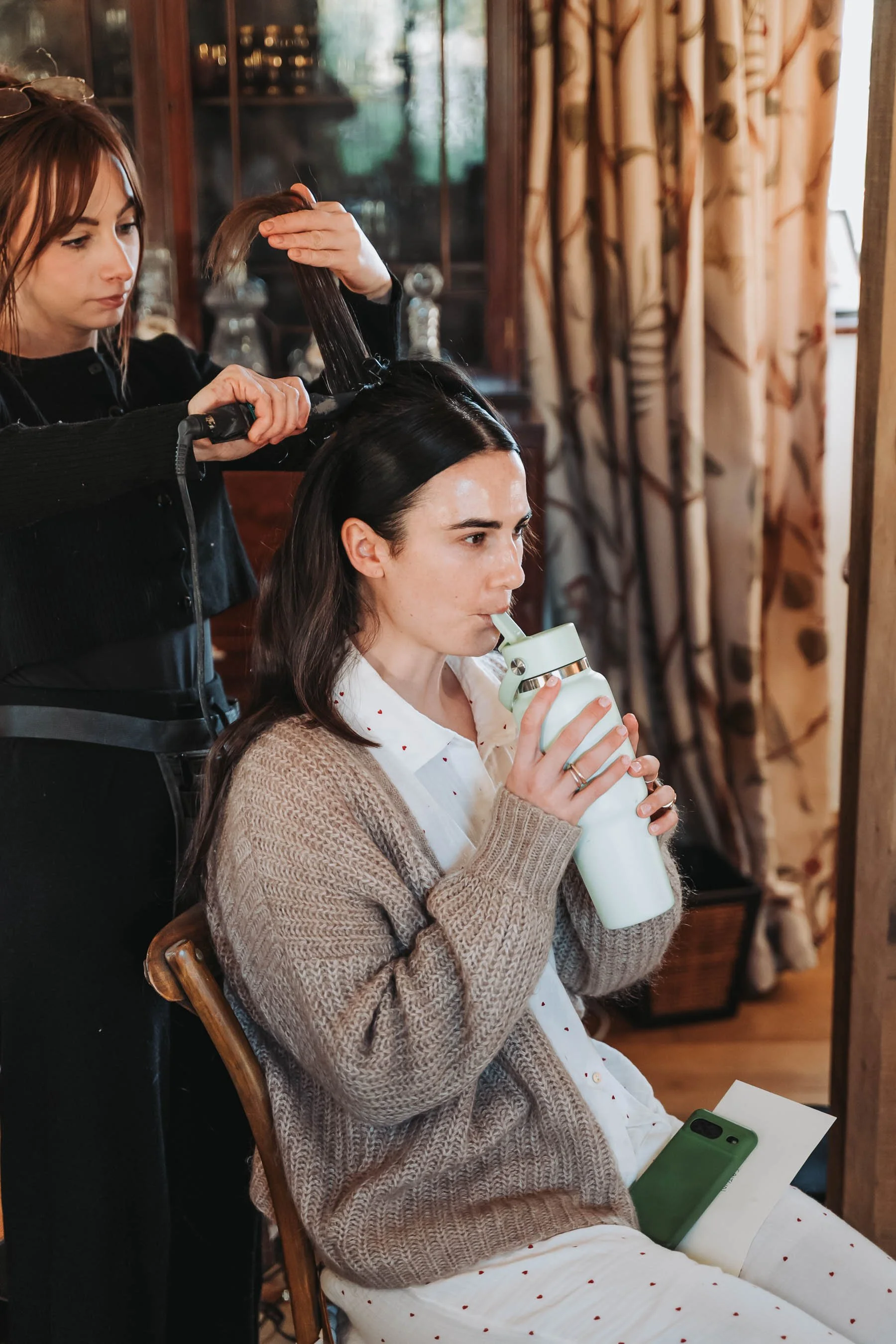 A woman sitting on a wooden chair drinking from a white tumbler with a green phone resting on her lap. A hairstylist is standing behind her, using a flat iron to style her dark hair in a cozy room with patterned curtains and a wooden cabinet.