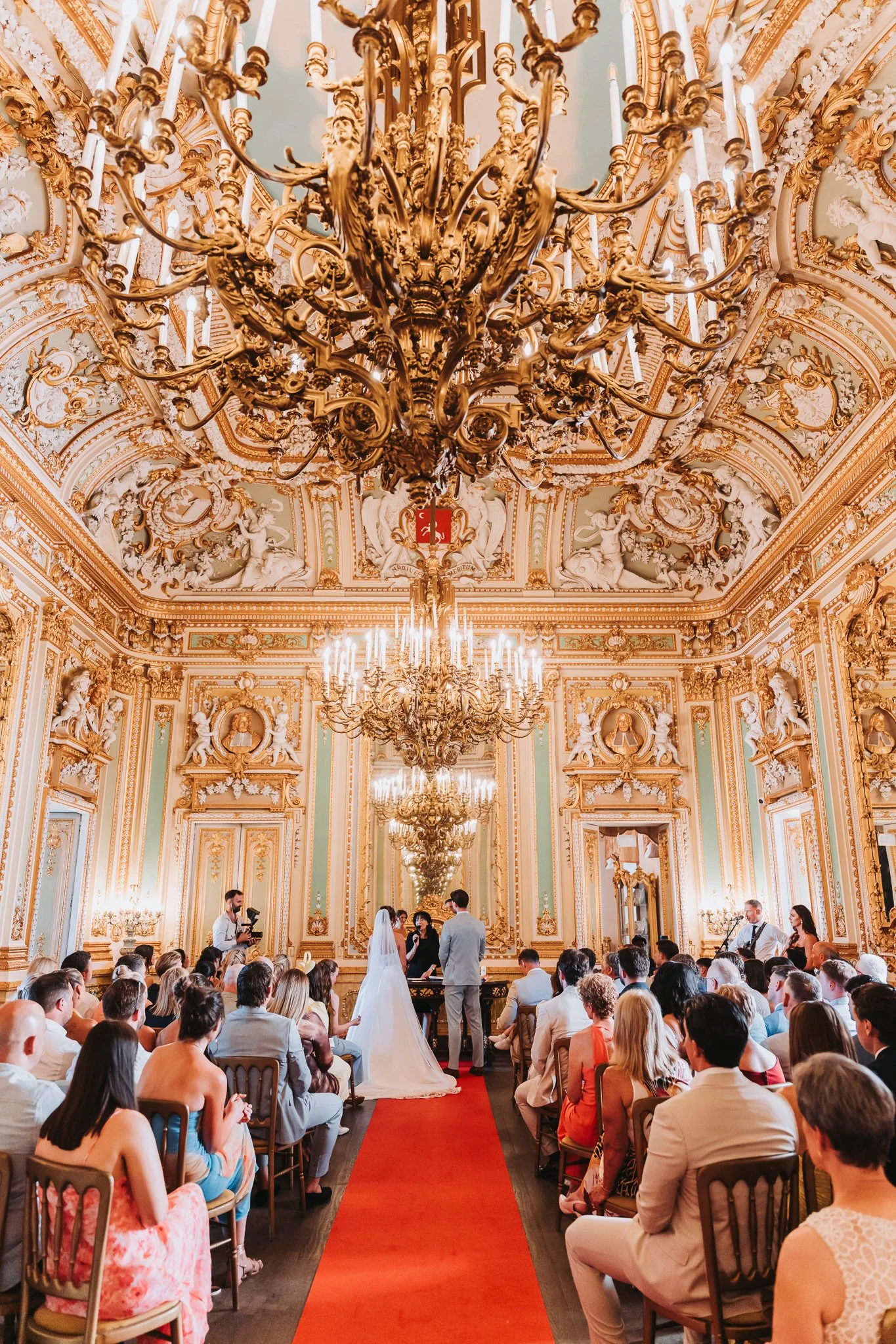 A wedding ceremony taking place in a grand, ornate room with a high ceiling and chandeliers. The bride and groom stand before an officiant, surrounded by seated guests. The room features gold detailing, sculpted walls, and classical artwork.