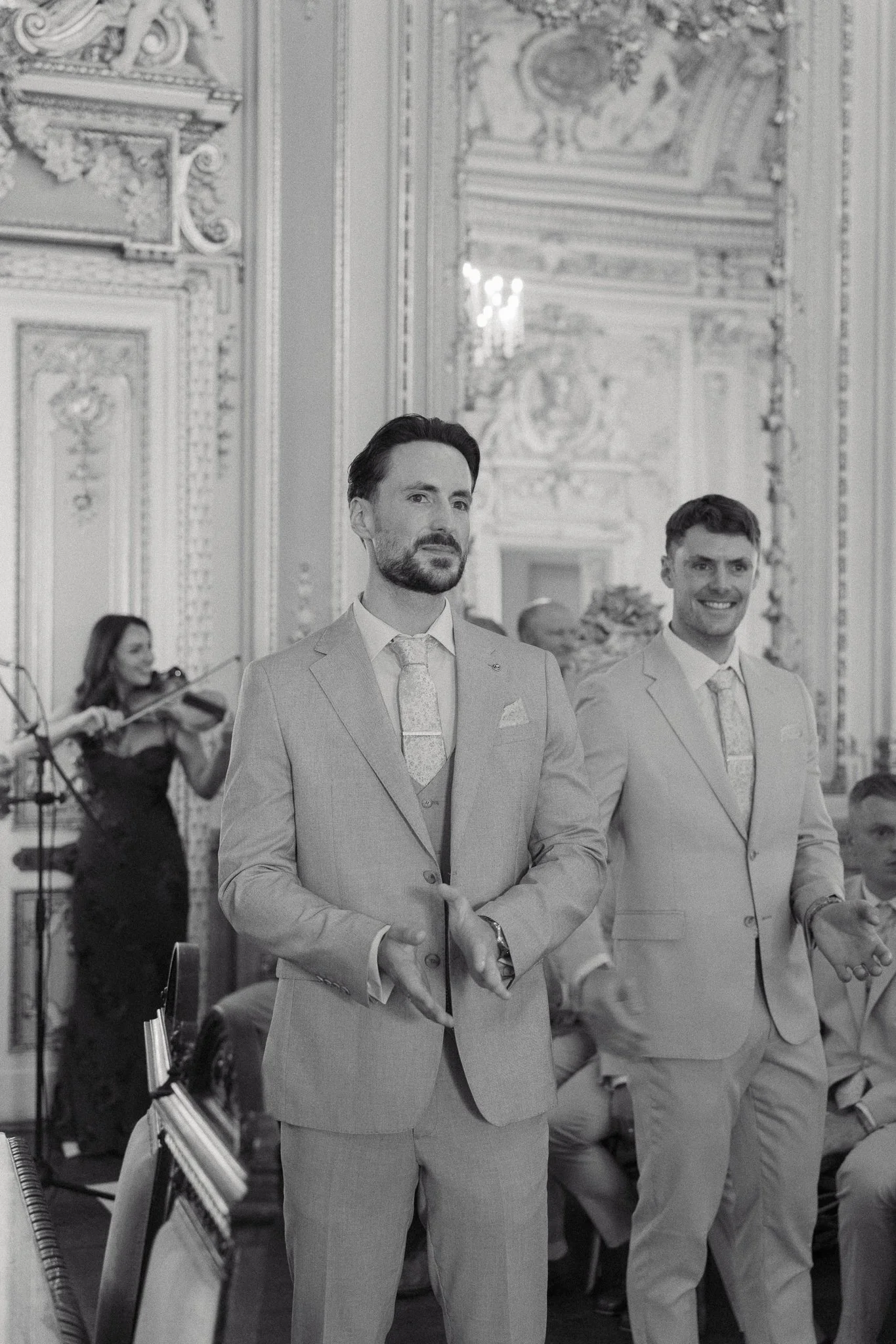 Two men in tuxedos standing in a decorated room, with a woman playing the violin in the background.