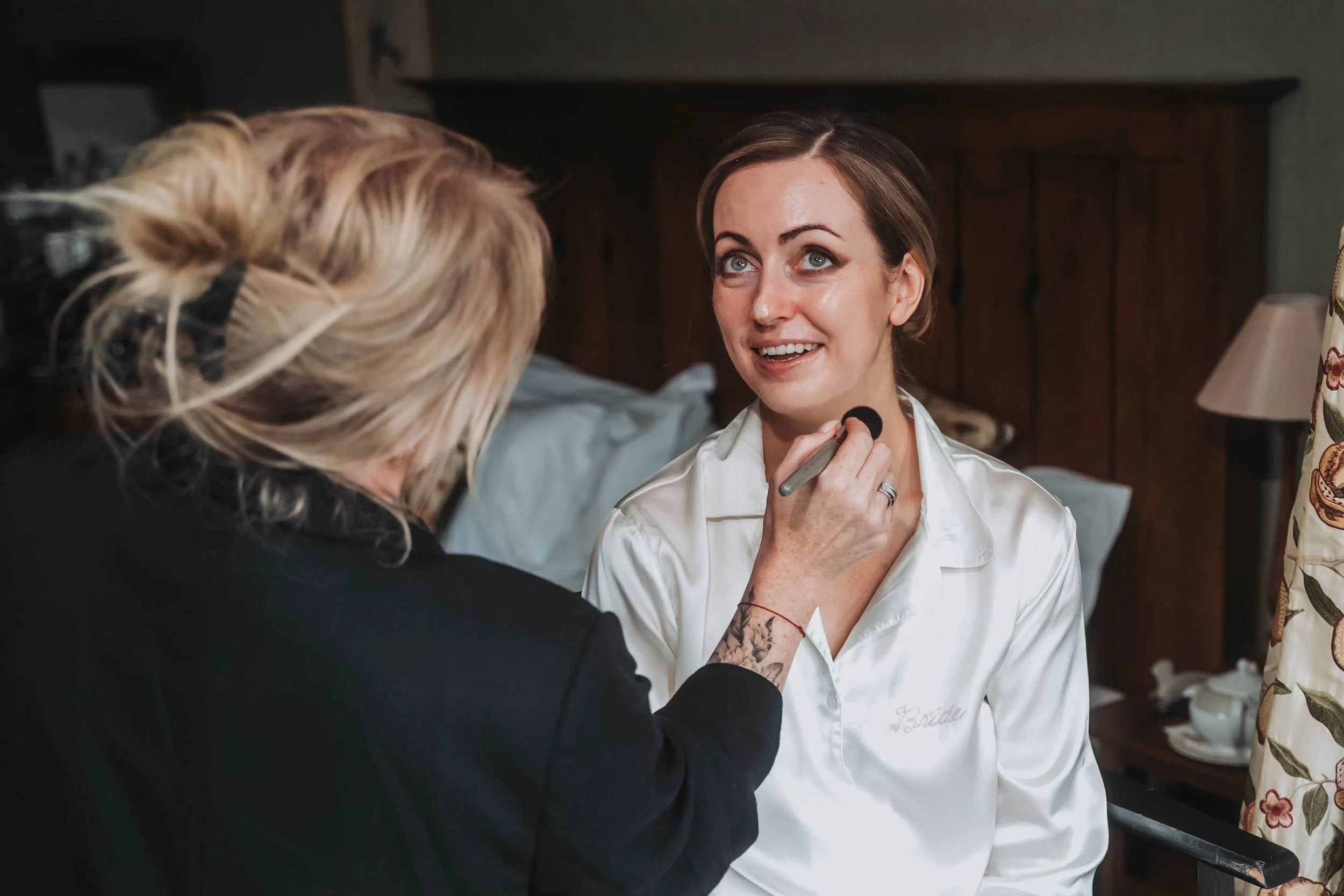 A woman with blue eyes and brown hair, wearing a white satin robe, is getting her makeup done by an artist with blonde hair and tattoos, in a cozy room with wooden furniture and floral curtains.