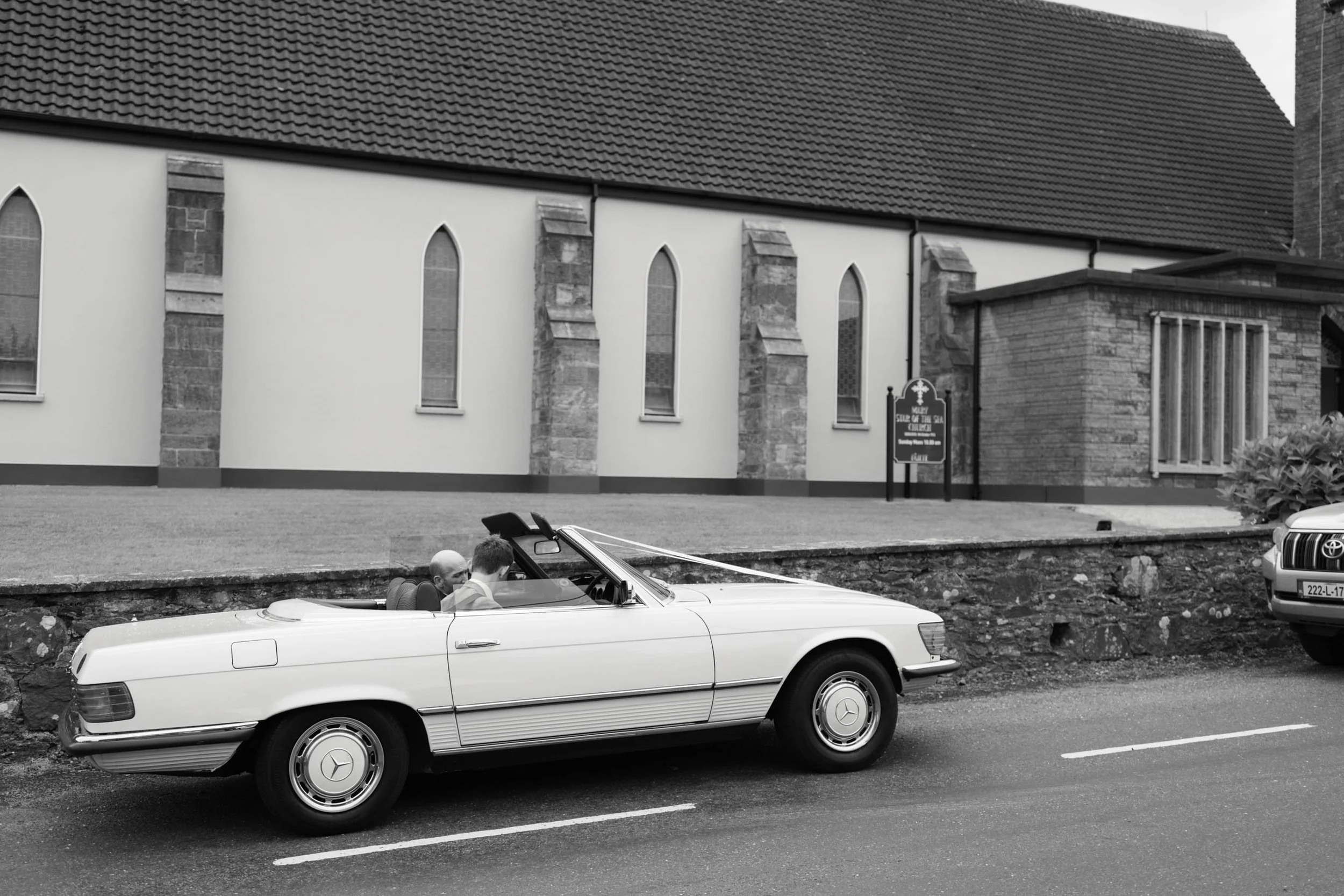 A black-and-white photo of a classic white convertible car with two people inside, driving on a street. Behind the car, there is a church with arched windows and a tiled roof, and a sign that reads 'MUIR CHURCH OF IRELAND - O'Connell'.