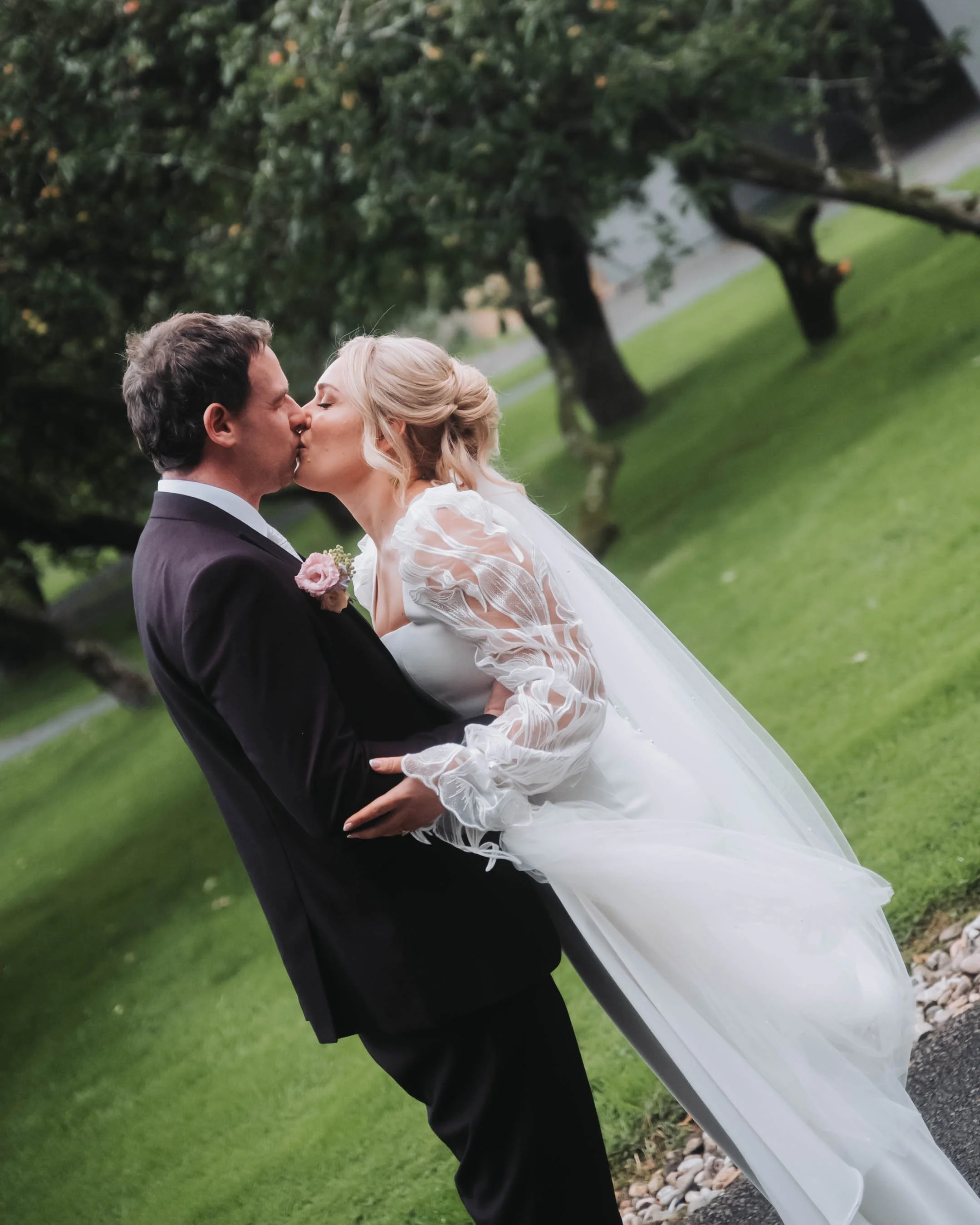 A newlywed couple, a man in a dark tuxedo and a woman in a white wedding dress, share a kiss outdoors on a sunny day, with green grass and trees in the background.
