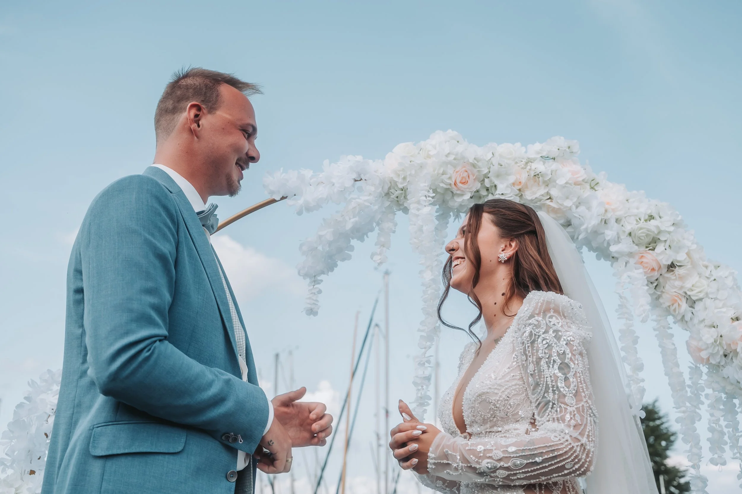 A bride and groom holding hands during their wedding ceremony, standing under a floral arch with white and blush roses, in an outdoor setting with a clear sky.