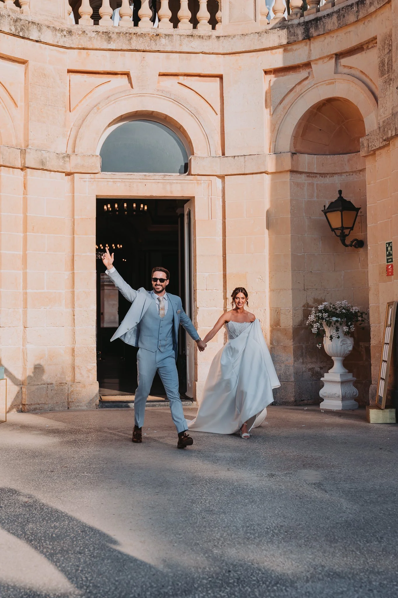A newlywed couple holding hands and walking out of a historic stone building, celebrating their wedding day. The man is dressed in a light gray suit, and the woman is wearing a white wedding gown. They are smiling and appear joyful.