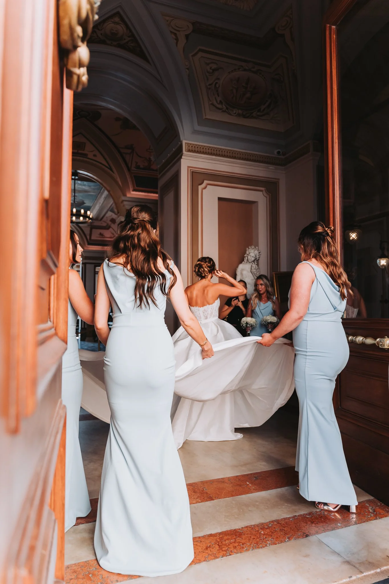 Bridesmaids in light blue dresses assisting a bride in a white wedding dress as she enters a room with ornate ceiling decor and a statue of a woman in the background.