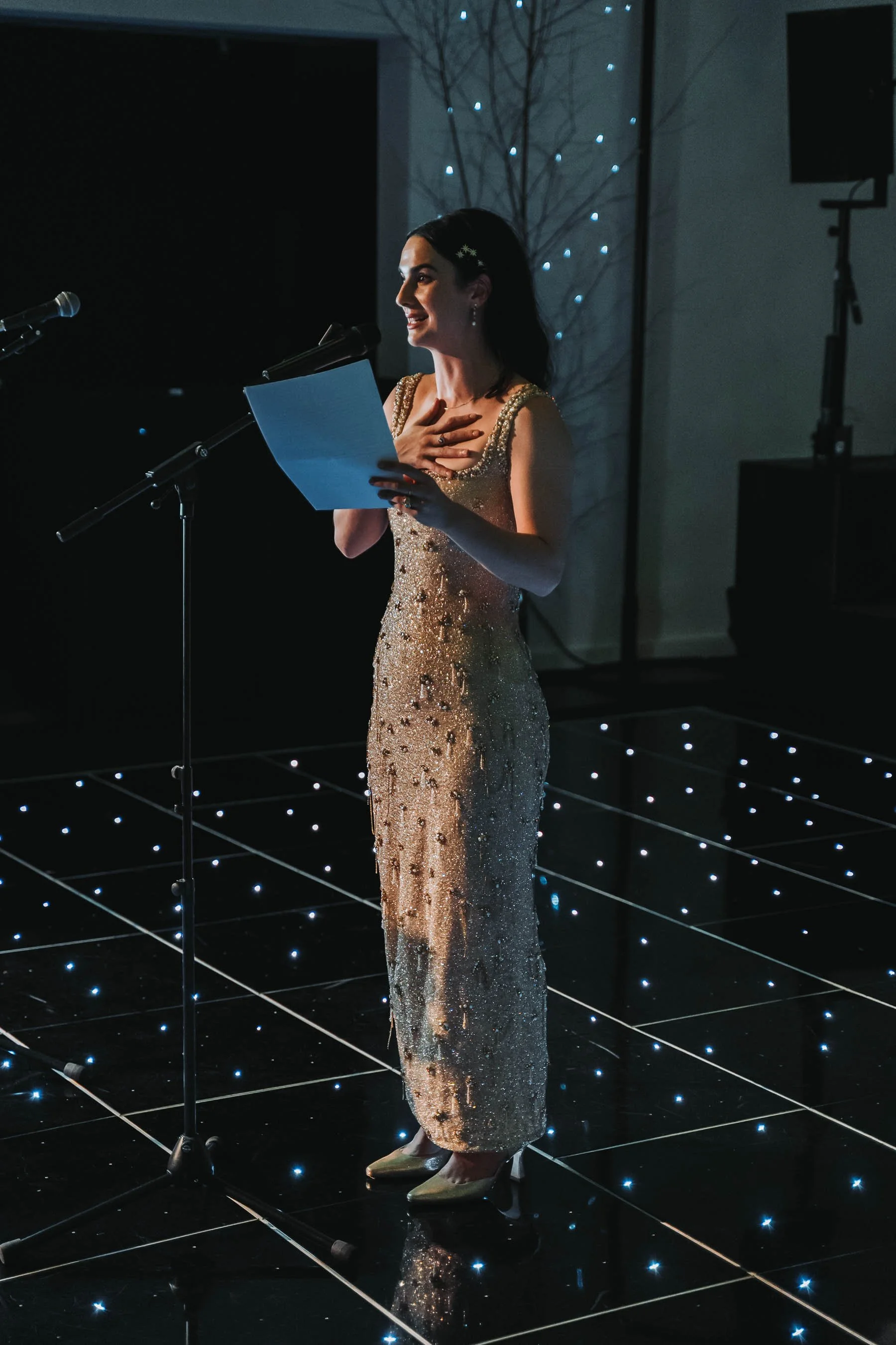 A woman in a shimmering gold gown with floral embellishments, standing in front of a microphone on a stage with black and white grid flooring illuminated by small white lights. She has dark hair styled with a hair clip and is expressing emotion, hold