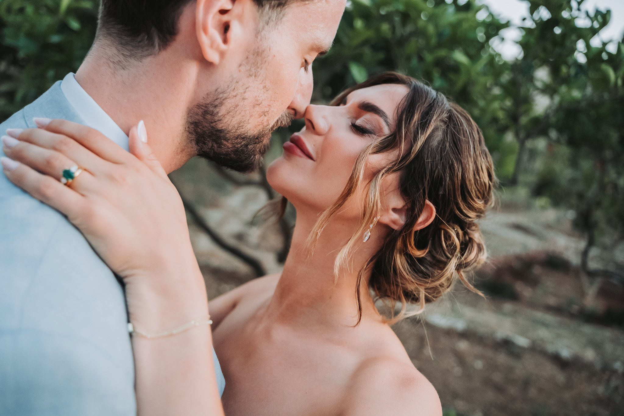 A close-up of a couple standing outdoors, about to kiss, with the woman holding the man's shoulder, trees in the background, and natural lighting.
