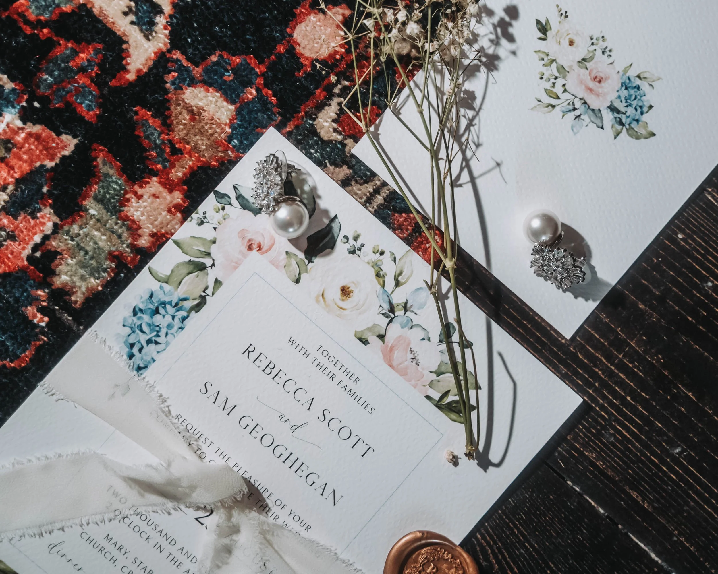 Wedding invitation with floral design, a pair of pearl earrings, and dried flowers on a dark wooden surface
