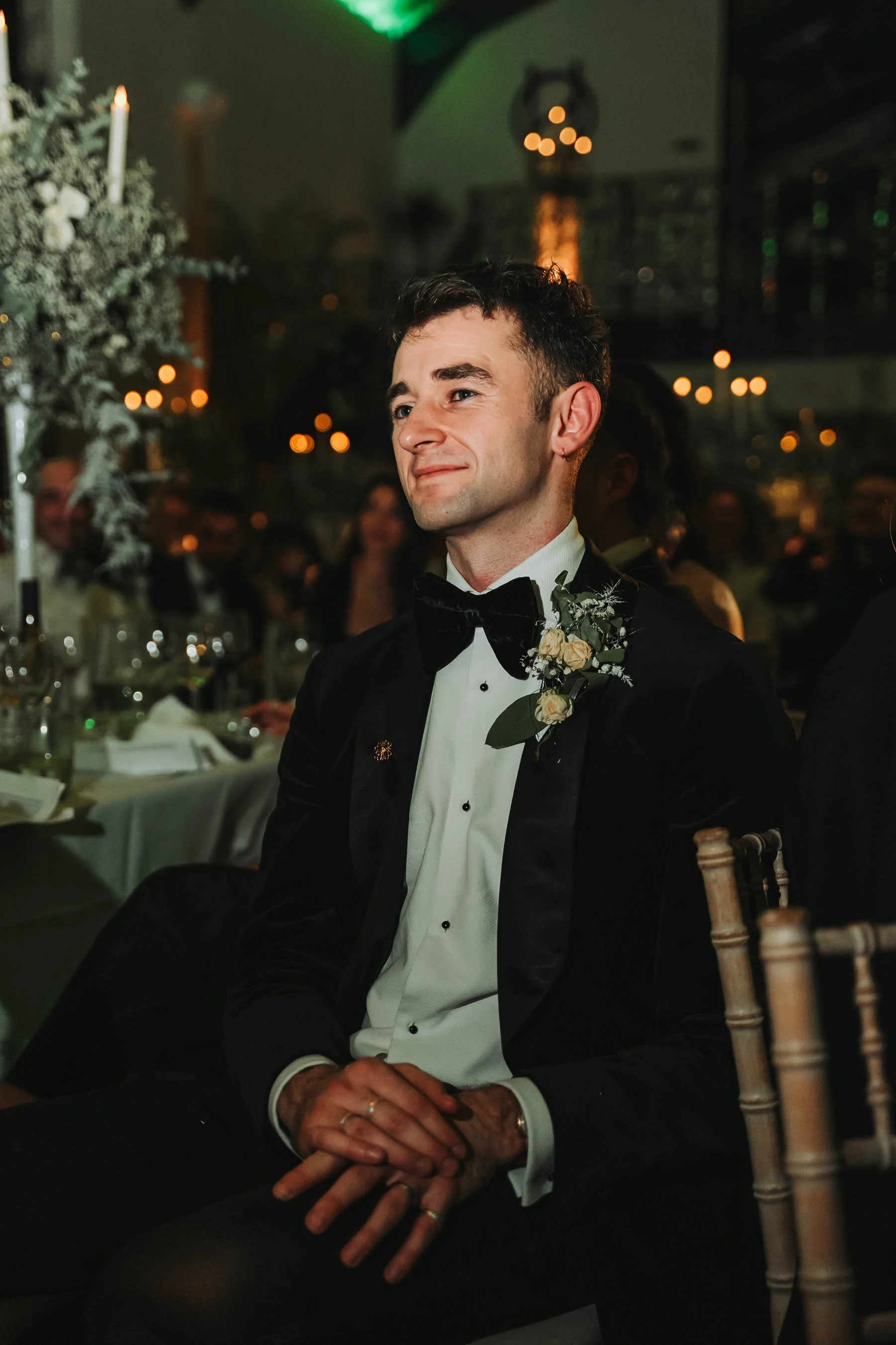 A young man with dark hair dressed in a tuxedo with a bow tie, sitting at a formal event. He has a small boutonniere on his lapel. The background shows a dark, elegant setting with blurred warm lights, floral arrangements, and guests in formal attire