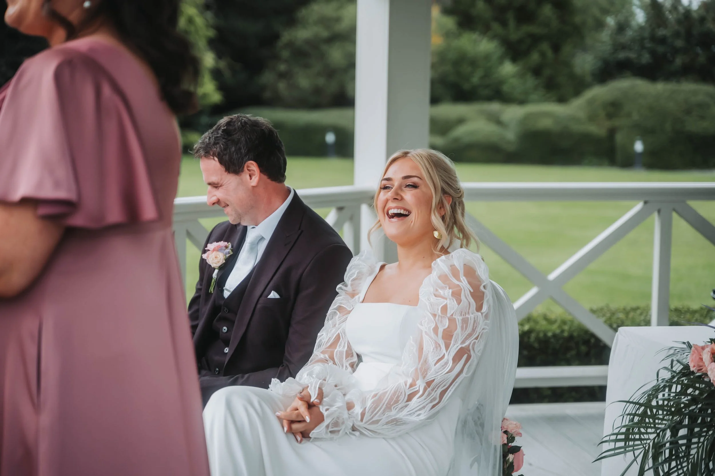 A bride and a groom sitting during a wedding ceremony outdoors, with the bride smiling and the groom looking down, while a woman in pink is standing in front of them.