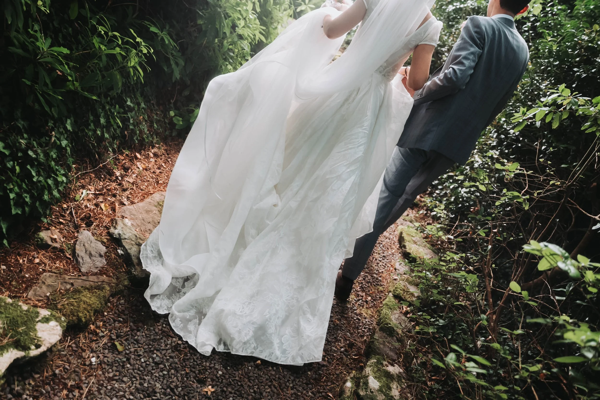 A bride in a white wedding dress walking on a dirt trail through a forest, holding hands with a groom in a gray suit.