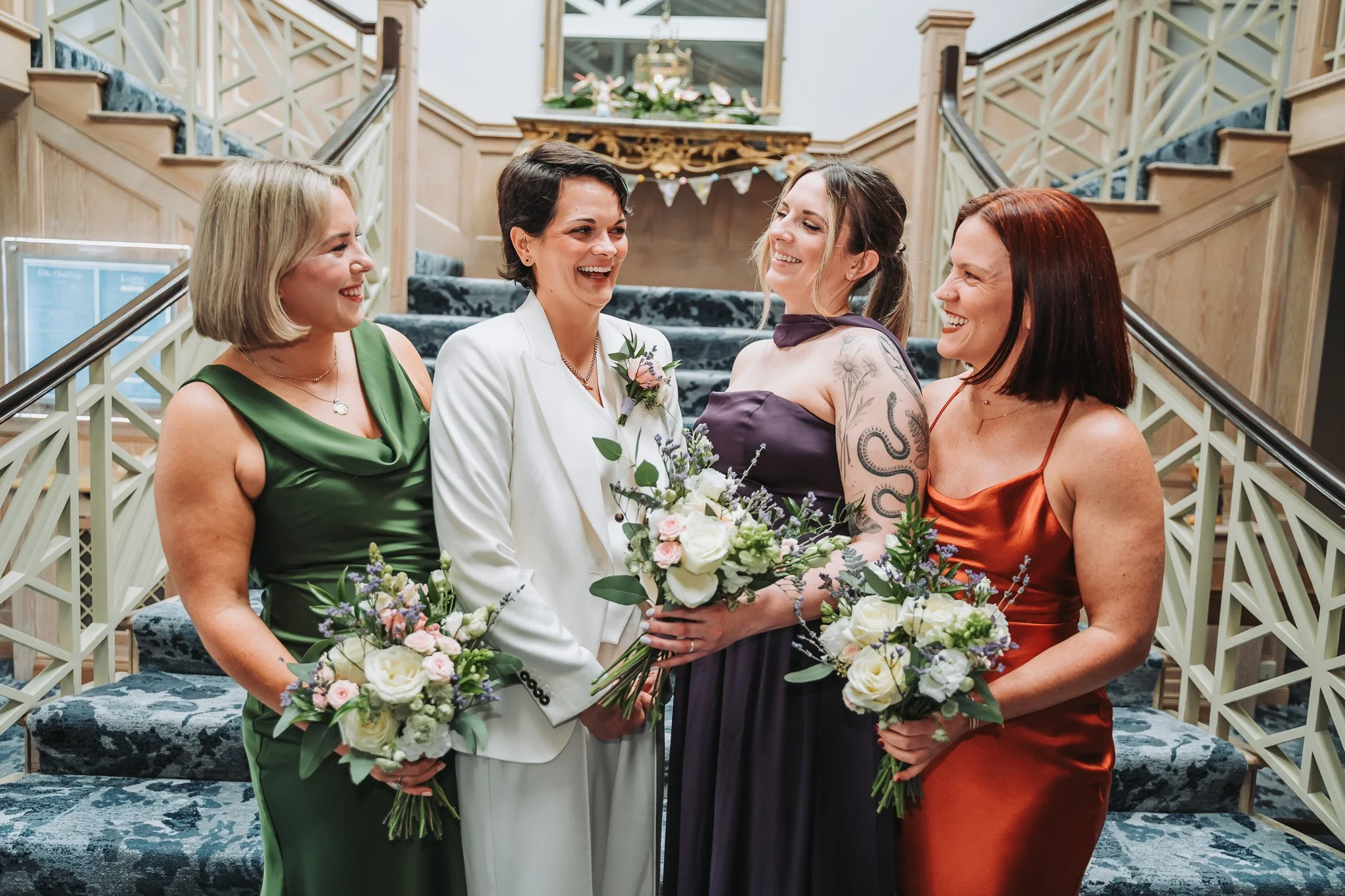 Four women standing on a staircase, smiling and holding bouquets of flowers, dressed in elegant attire, with a decorative railing and floral garland in the background.