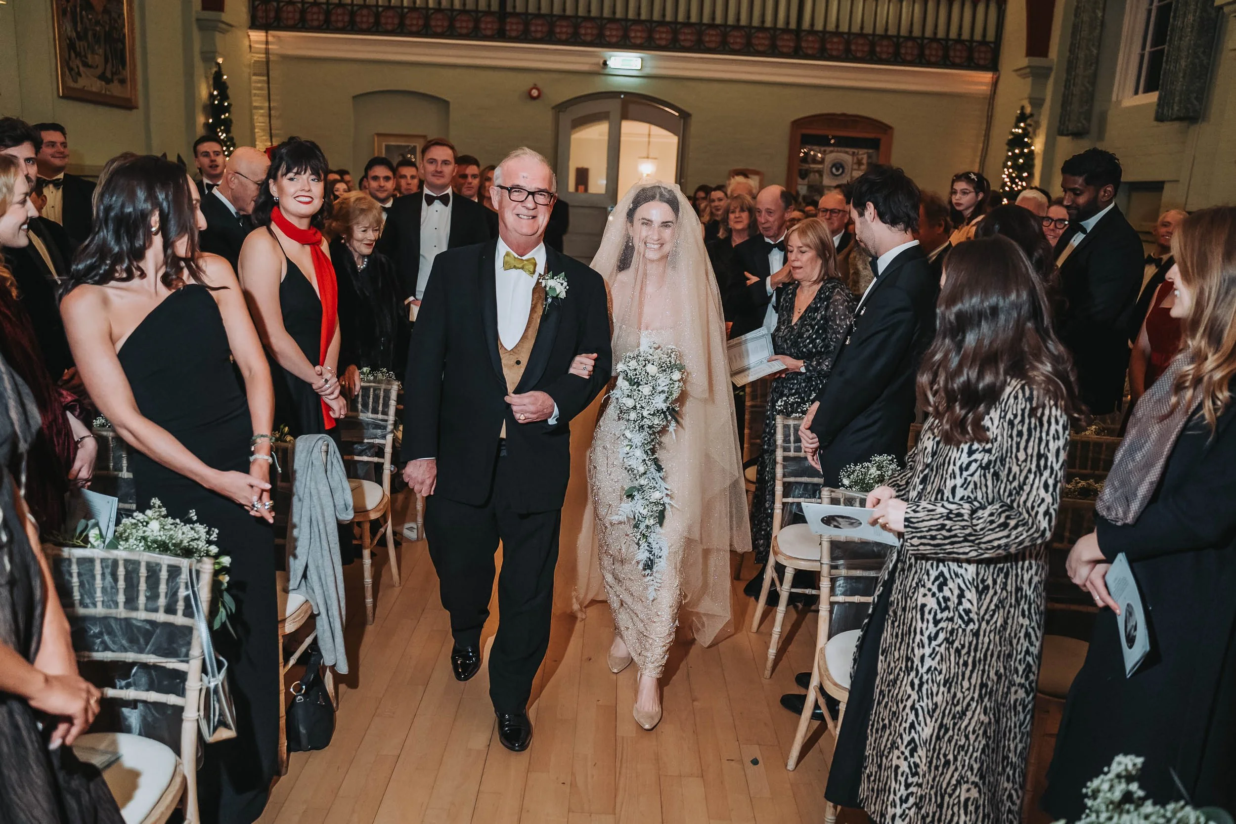 A wedding ceremony with the bride walking down the aisle accompanied by an older man, possibly her father, in a formal indoor setting with guests dressed in elegant attire.