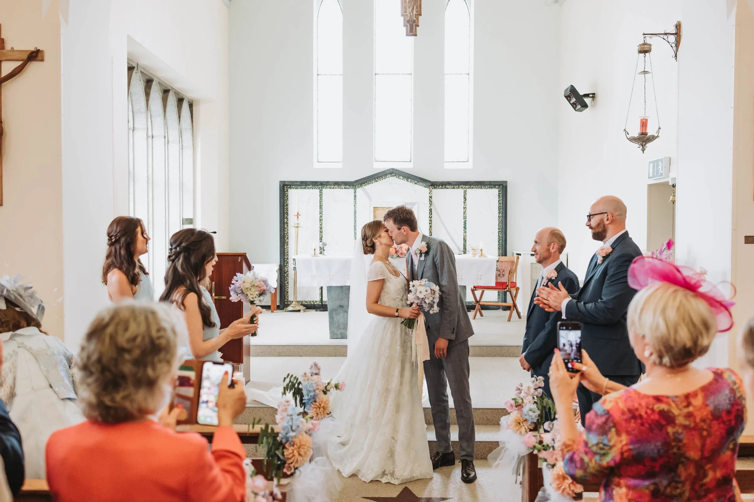 A wedding ceremony inside a church with a bride and groom kissing. The bride is holding a bouquet, and the groom is wearing a gray suit with a pink boutonniere. Guests are seated on both sides, some taking photos. The church features tall windows, a 