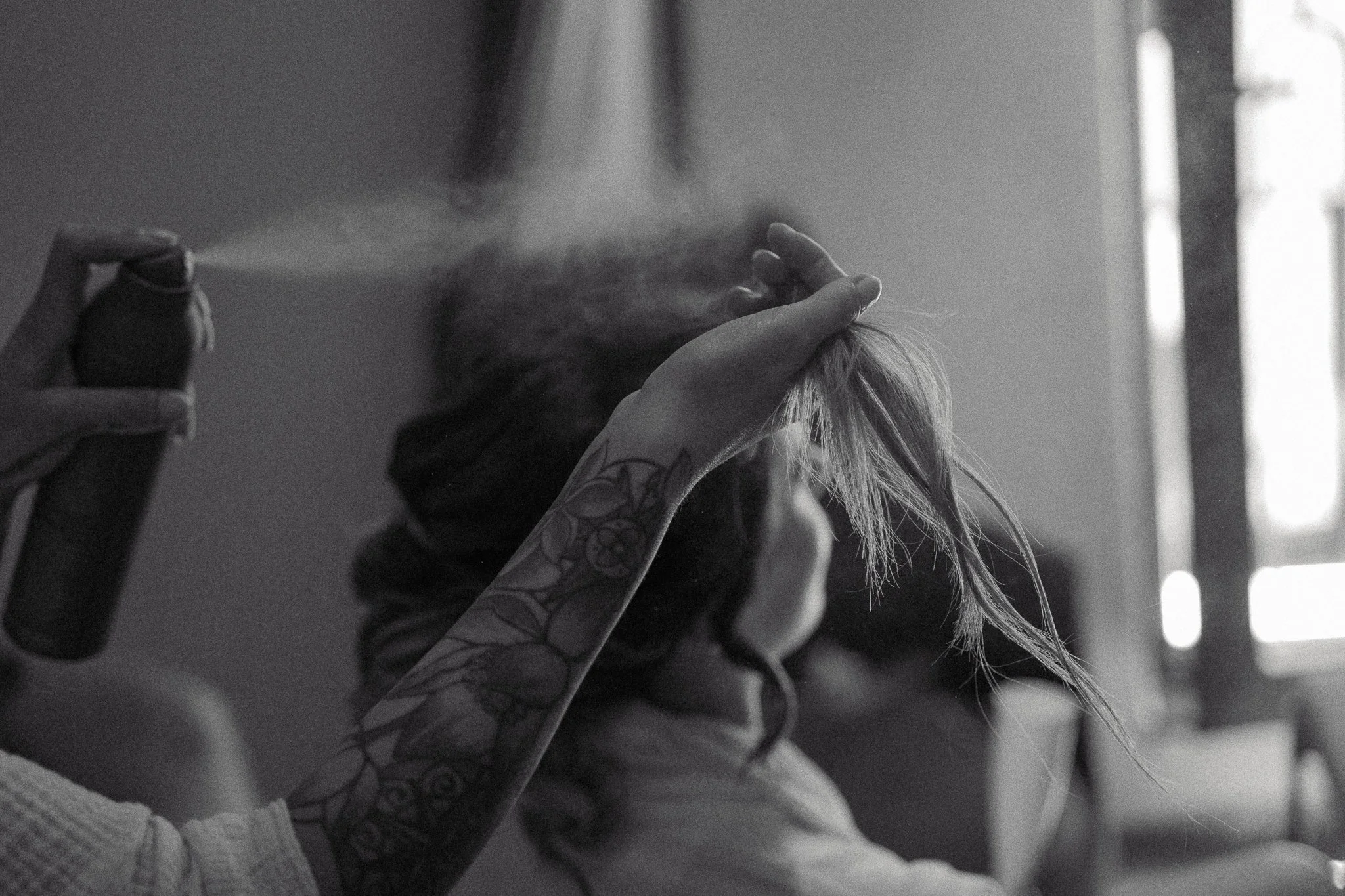 A person with tattoos on their arm is smoking a cigarette or joint, with smoke visible near their face, in a room with bright natural light coming from a window.
