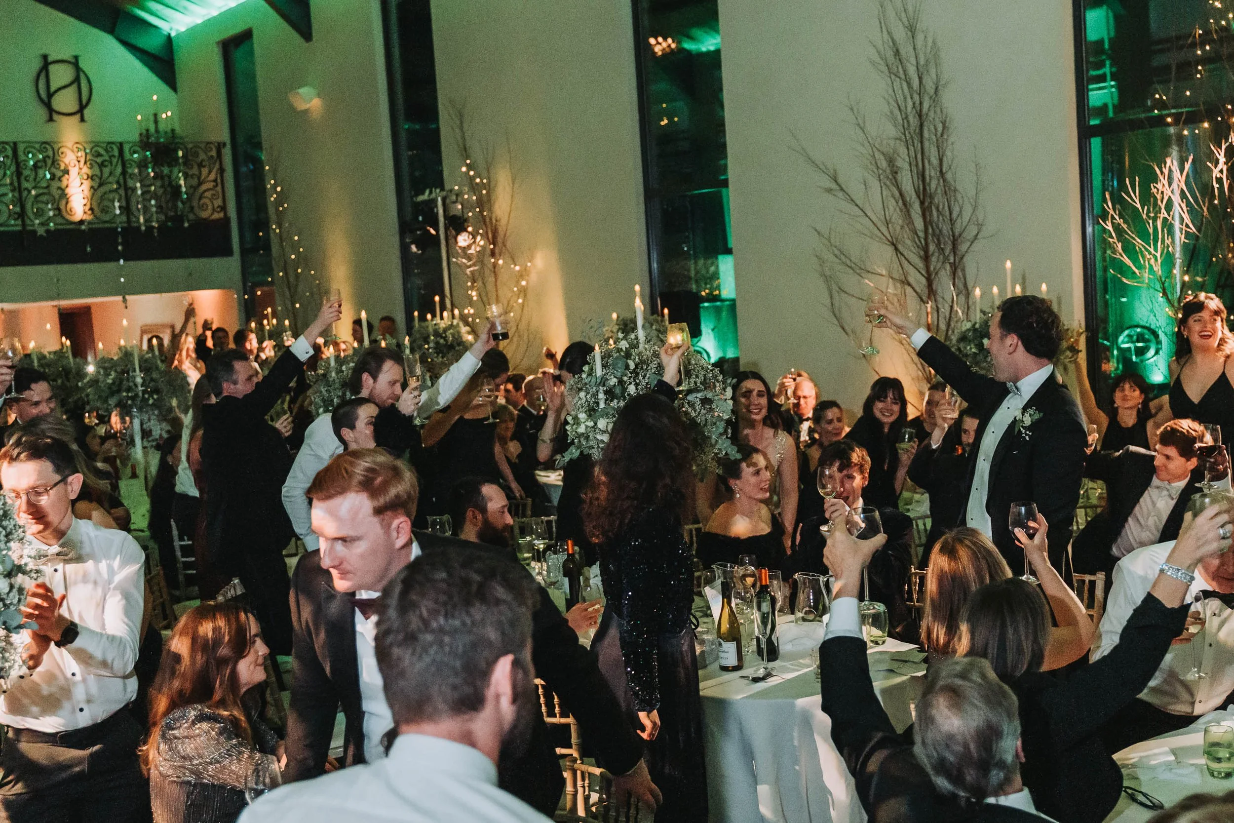 A large group of people dressed in formal attire at a wedding reception or celebration, raising glasses in toast, with a decorated venue featuring candles, floral arrangements, and tall tree branches in the background.
