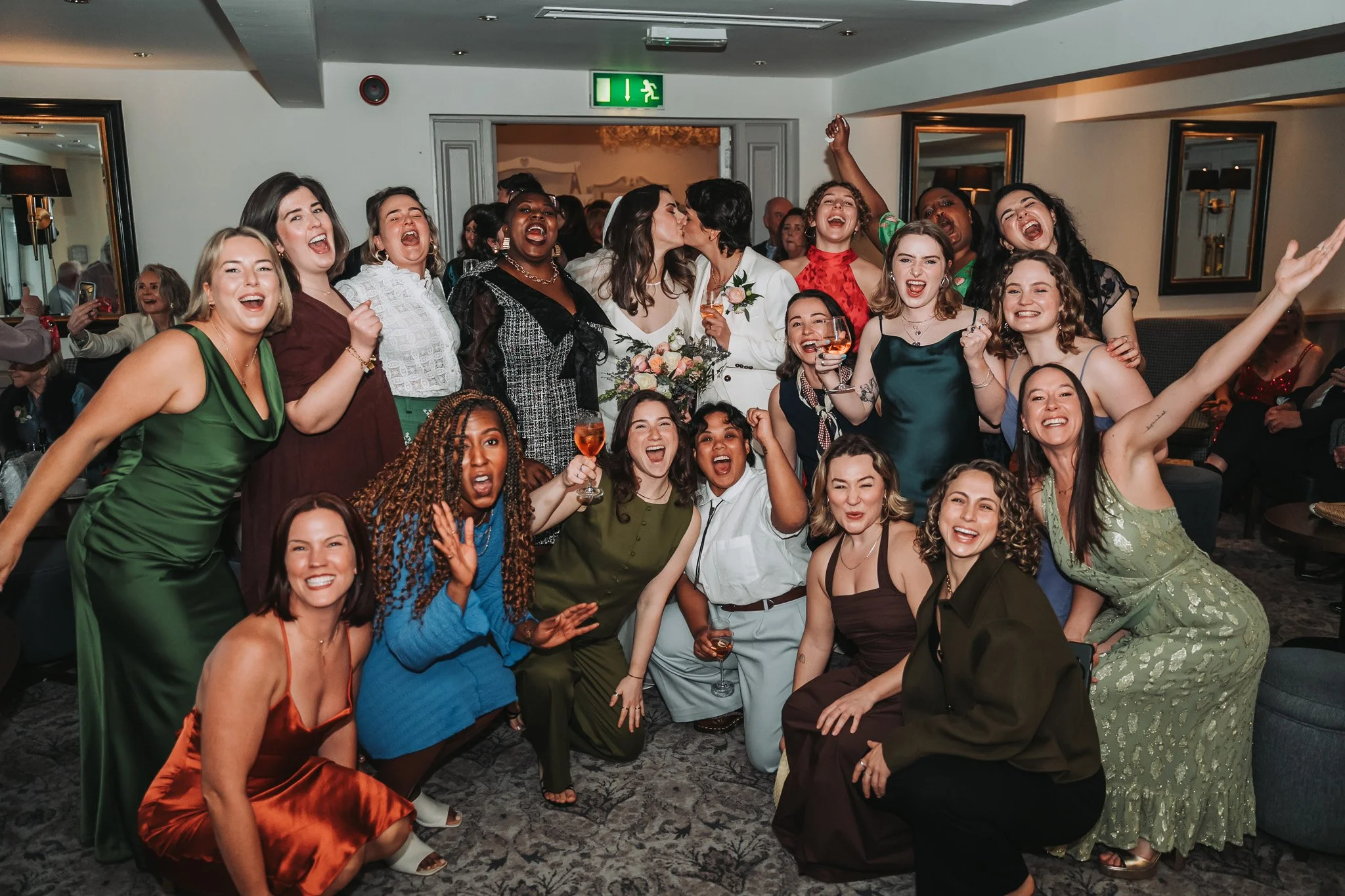 A large group of diverse women celebrating and posing together at a party, some holding glasses of rosé wine, indoors with mirrors and wall lamps.